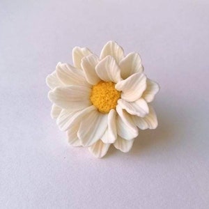 May include: A white daisy flower with a yellow center. The flower is made of polymer clay and is a handmade craft.