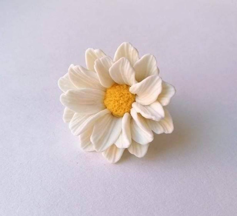 May include: A white daisy flower with a yellow center. The flower is made of polymer clay and is a handmade craft.