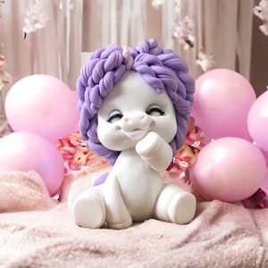 Könnte beinhalten: Eine weiße Einhorn-Figur mit einer lila geflochtenen Mähne und einem Schwanz. Das Einhorn sitzt mit erhobenem rechten Bein und geneigtem Kopf. Die Figur ist von rosa Luftballons umgeben.