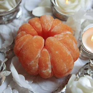 Peut inclure: Gros plan sur une bougie ronde de couleur orange vif, texturée, en forme d'agrume. La bougie est posée sur une surface blanche avec des objets décoratifs blancs et argentés.