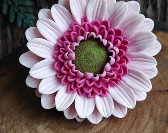 May include: A pink and white gerbera daisy with a green center. The flower is made of polymer clay and is a realistic representation of a real flower.