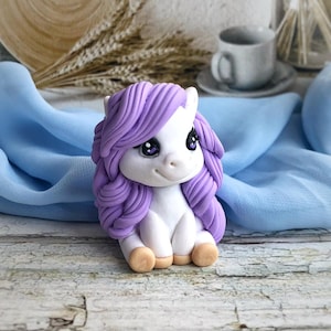May include: A small white unicorn figurine with purple hair, sitting on a white wooden surface. The unicorn has large, blue eyes and a sweet expression.
