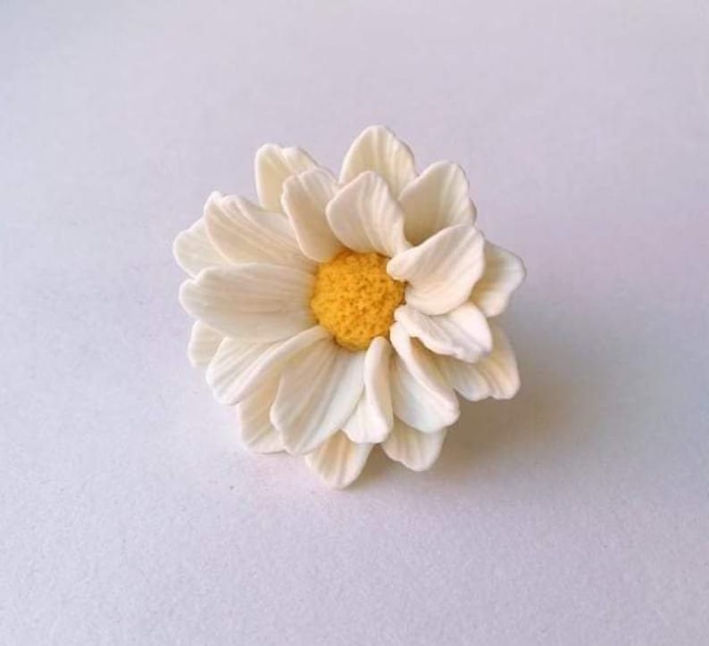May include: A white daisy flower with a yellow center. The flower is made of polymer clay and is a delicate and realistic representation of a daisy.