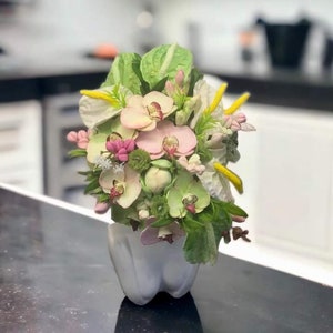 Könnte beinhalten: Ein Strauß aus rosa und weißen Orchideen, weißen Anthurienblüten und grünem Laub, arrangiert in einer weißen Keramikvase in Zahnform.