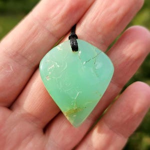 May include: A light green gemstone pendant with a black cord. The pendant is a unique, shield-like shape with natural inclusions and a polished surface. The gemstone is held in a hand against a blurred green background.