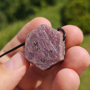 Könnte beinhalten: Ein facettierter, roher Rubinanhänger mit schwarzem Band. Der Edelstein hat eine tiefrosa-rote Farbe mit dunklen Einschlüssen. Der Anhänger wird in einer Hand vor einem verschwommenen grünen Hintergrund gehalten.