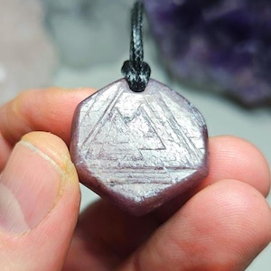 May include: A hexagonal pendant with a mountain design carved into its surface. The pendant is a light purple colour and is attached to a black cord. The pendant is held by a person's hand.
