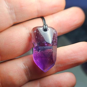 May include: A faceted amethyst pendant with a dark purple hue, suspended from a black cord. The crystal is held in a hand, showcasing its polished surface and natural variations. The pendant has a pointed bottom and a hole at the top for the cord.