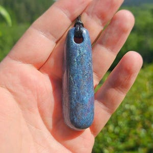 May include: A polished, elongated blue pendant with a smooth, glossy finish and subtle reddish-purple mineral inclusions. The stone features a drilled hole at the top, through which a thin black cord is threaded for suspension as a necklace.
