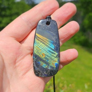 Peut inclure: Un pendentif en labradorite poli de forme rectangulaire aux coins arrondis, présentant une pierre de base gris foncé avec des éclats irisés vibrants de jaune, d'or et de bleu sur sa surface, suspendu à un simple collier en cordon noir.