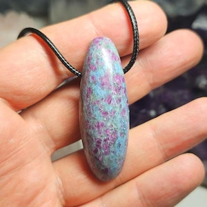 May include: A gemstone pendant necklace. The polished, oval-shaped stone has blue and purple hues, suspended from a black cord. The pendant is held in a hand, showing its size and colour variations. The stone's length is approximately 4 cm.