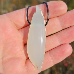 Può includere: Una collana con pendente allungato e pallido con un cordino nero. Il pendente è di un colore chiaro e traslucido, forse giada o una pietra simile. La forma è irregolare, con una superficie liscia e lucida.
