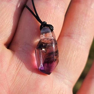 May include: A faceted, teardrop-shaped pendant necklace featuring a translucent purple and brown crystal. The crystal is suspended from a black cord, creating a simple and elegant design. The pendant is held in a hand, showcasing its natural beauty.