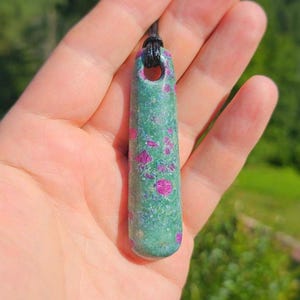 Puede incluir: Un colgante cilíndrico alargado de piedra verde con inclusiones cristalinas de color rosa brillante. El colgante presenta un acabado liso y pulido, con un orificio en la parte superior por el que pasa un cordón negro fino para colgarlo.