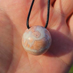 Puede incluir: Un collar con colgante redondo con una piedra pulida que muestra un patrón veteado de tonos blancos y melocotón. La piedra cuelga de un cordón negro. El colgante es una piedra natural, probablemente una gema.