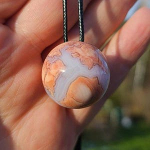 May include: A spherical pendant with a marbled design in orange, white, and brown, hanging from a black cord. The pendant's surface has a natural, stone-like appearance. The background is out of focus.