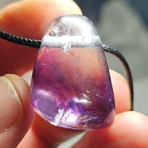 May include: A polished amethyst pendant with a black cord. The gemstone is a mix of clear, light pink, and deep purple hues. The pendant is held in a person's hand, showcasing its smooth, rounded shape and natural color variations.