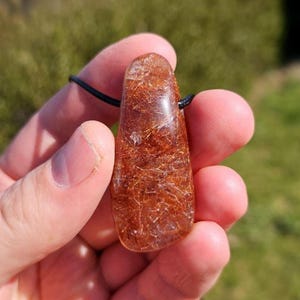 May include: A teardrop-shaped pendant made of clear quartz with inclusions of red copper. The pendant is on a black cord.