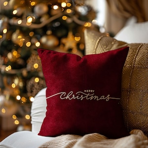 Puede incluir: Un cojín cuadrado de color burdeos con las palabras "Merry Christmas" en elegante escritura blanca. El cojín está colocado sobre una superficie blanca, con un cojín dorado estampado y un árbol de Navidad al fondo.
