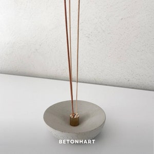 Könnte beinhalten: Grauer Beton-Räucherstäbchenhalter mit Messing-Einsatz, der drei Räucherstäbchen hält. Der Halter hat eine flache, schalenartige Form. Der Text "BETONHART" ist auf der Unterseite aufgedruckt.