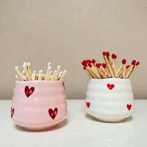 May include: Two ceramic match holders, one pink and one white, each decorated with red heart designs. The pink holder contains white-tipped matches, while the white holder has red-tipped matches. Both are on a white surface.