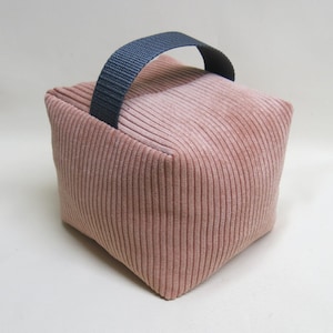Könnte beinhalten: Ein türstopperförmiger Würfel aus rosa Cordstoff. Es hat eine vertikale Rippenstruktur und einen marineblauen Stoffgriff an der Oberseite. Der Türstopper ist dazu bestimmt, Türen offen zu halten oder zu verhindern, dass sie zuschlagen.