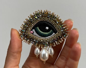 May include: A green eye brooch with gold and iridescent bead embellishments and a dangling pearl drop.