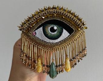 May include: Embroidered eye-shaped brooch with a detailed design. The eye features a green iris and is surrounded by gold-colored beads and fringe. The brooch has a decorative, ornate style.