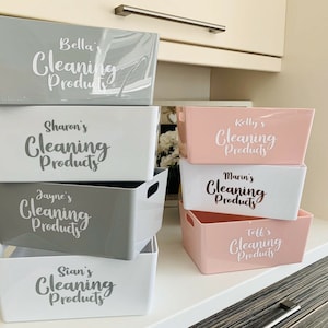 May include: Stack of five grey and pink plastic storage bins with white lettering. The bins are labelled with names and the words "Cleaning Products".