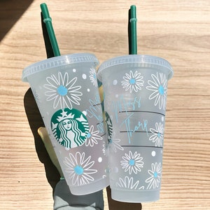 May include: Two clear plastic cups with green straws. The cups have a white daisy pattern with blue centres. One cup has the text "Miss Thorpe" written on it. The other cup has a Starbucks logo.