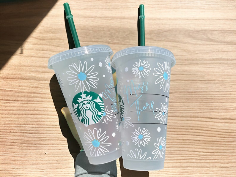 May include: Two clear plastic cups with green straws. The cups have a white daisy pattern with blue centres. One cup has the text "Miss Thorpe" written on it. The other cup has a Starbucks logo.