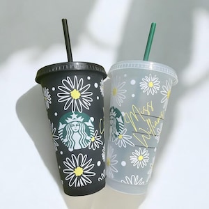 May include: Two reusable Starbucks cups with black and white daisy patterns. One cup is black with a Starbucks logo and the other is clear with the text "Miss Williams".