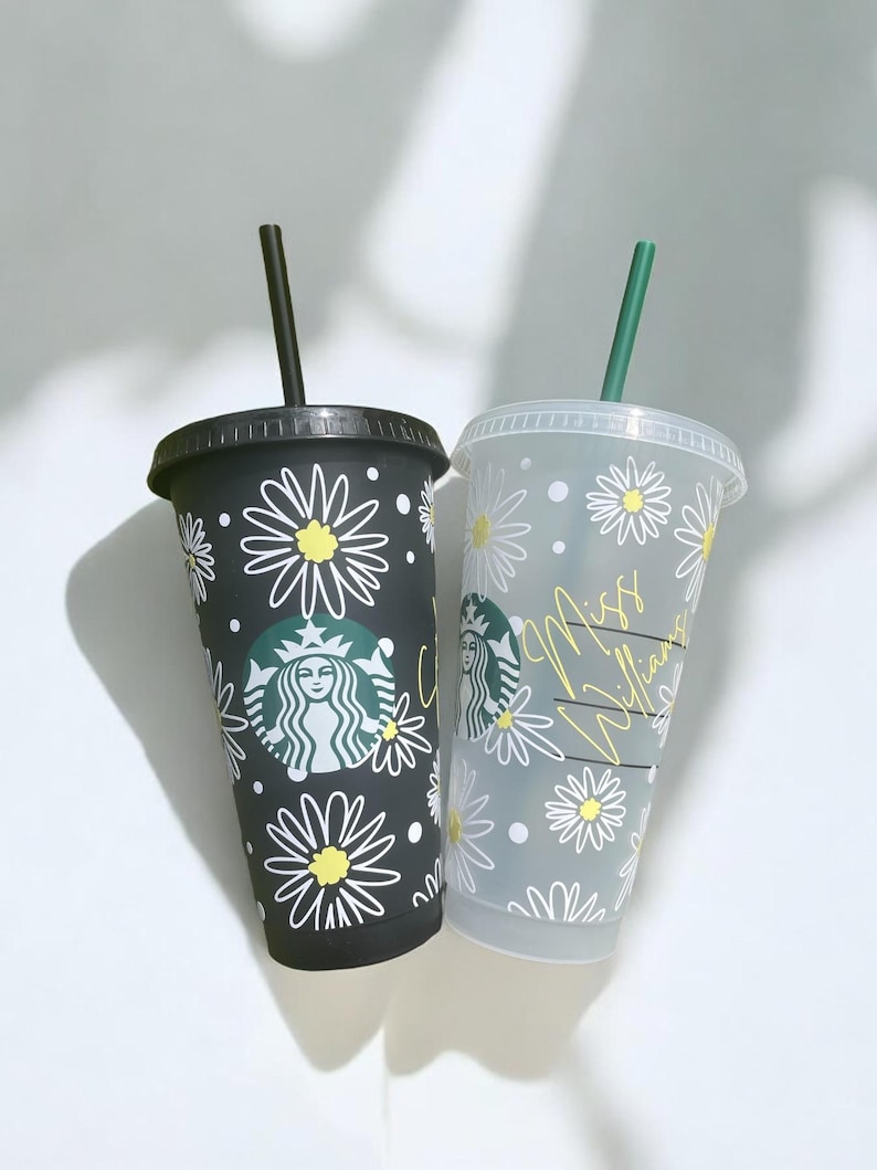 May include: Two reusable Starbucks cups with black and white daisy patterns. One cup is black with a Starbucks logo and the other is clear with the text "Miss Williams".