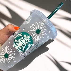 May include: A white reusable cup with a green Starbucks logo and a white daisy pattern. The cup has a green straw.
