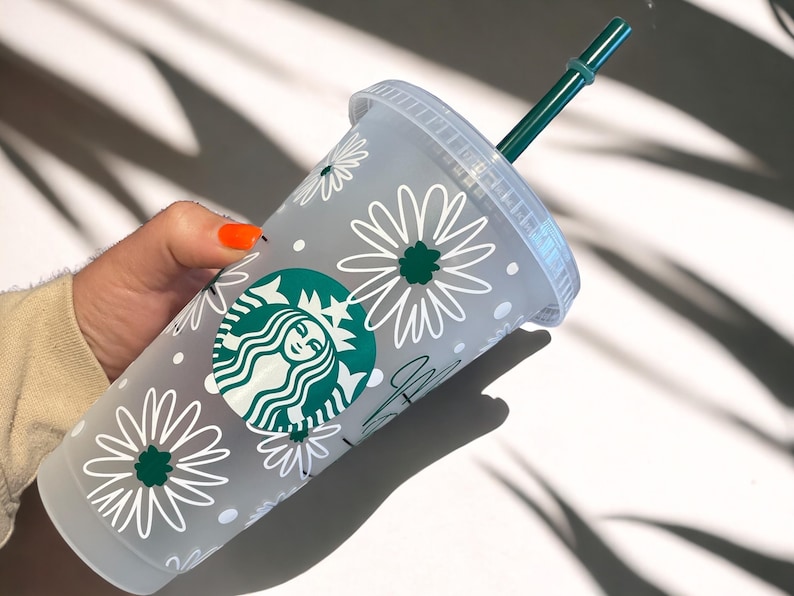 May include: A white reusable cup with a green Starbucks logo and a white daisy pattern. The cup has a green straw.