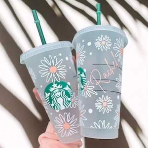 May include: Two clear plastic cups with lids and straws. The cups are decorated with a white daisy pattern and pink accents. One cup has the Starbucks logo and the other has the name "Rachel" written on it.