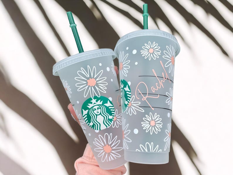 May include: Two clear plastic cups with lids and straws. The cups are decorated with a white daisy pattern and pink accents. One cup has the Starbucks logo and the other has the name "Rachel" written on it.