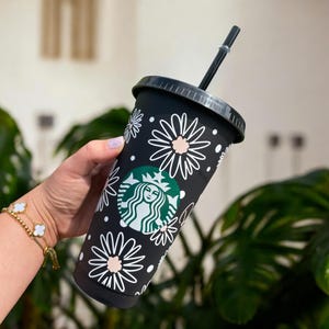 May include: Black reusable cup with a white daisy pattern and a green Starbucks logo. The cup has a black lid and a black straw.