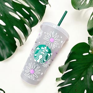 May include: A grey reusable Starbucks cup with a white daisy pattern and a green Starbucks logo. The cup has a green straw.