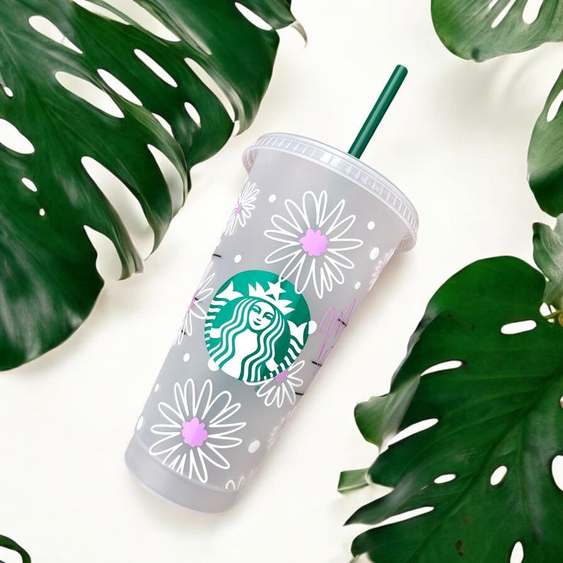 May include: A grey reusable Starbucks cup with a white daisy pattern and a green Starbucks logo. The cup has a green straw.