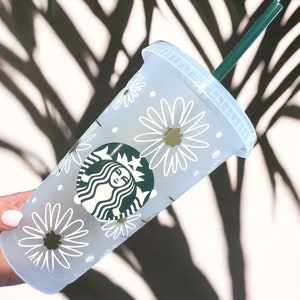 May include: White reusable cup with a green Starbucks logo and a white daisy pattern. The cup has a green straw.