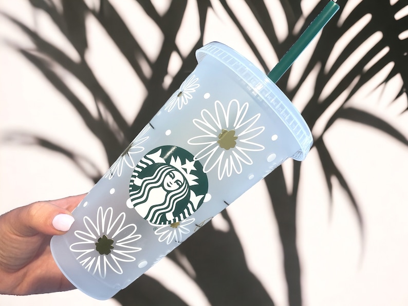May include: White reusable cup with a green Starbucks logo and a white daisy pattern. The cup has a green straw.