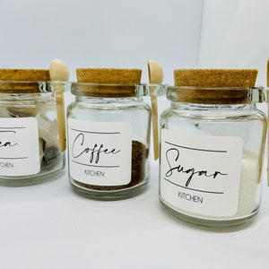 May include: Three glass jars with cork lids and wooden spoons. The jars are labelled "Tea", "Coffee", and "Sugar" with the word "Kitchen" underneath each label.