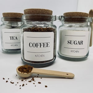 May include: Three glass jars with cork lids labelled "TEA", "COFFEE", and "SUGAR" with the word "KITCHEN" below each. A small wooden spoon rests near the jars, with coffee granules scattered around it. A minimalist kitchen storage set.