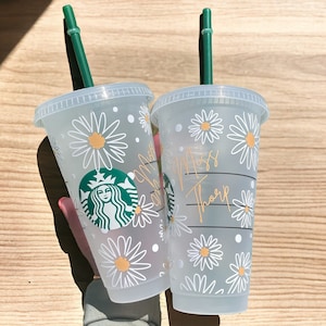 May include: Two clear plastic cups with green straws and white daisy designs. The cups have the text "Miss Tharp" and a Starbucks logo on them.