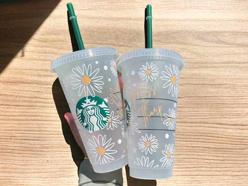 May include: Two clear plastic cups with green straws and white daisy designs. The cups have the text "Miss Tharp" and a Starbucks logo on them.