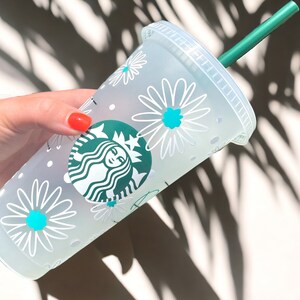 May include: A white reusable cup with a green Starbucks logo and a white daisy pattern. The cup has a green straw.