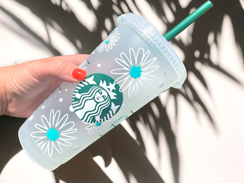 May include: A white reusable cup with a green Starbucks logo and a white daisy pattern. The cup has a green straw.