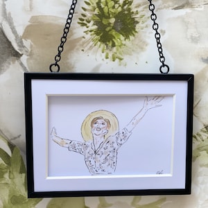 May include: A black framed print of a woman wearing a straw hat and a floral patterned shirt, with her arms raised in the air. The print is hanging on a wall with a floral pattern.