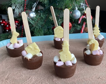 May include: Several chocolate cups filled with marshmallows and topped with white chocolate shapes, including a snowman, Santa, and a Christmas tree. Each cup has a wooden spoon. A Christmas tree is in the background.